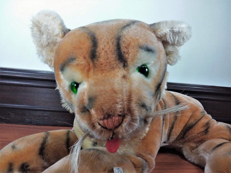 May include: A plush tiger toy with brown and black stripes, green eyes, and a pink tongue sticking out. The toy has a white mustache and is lying on a wooden floor.
