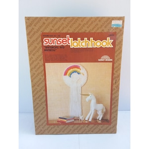 May include: A Sunset Designs latch hook kit box titled Raindrops and Rainbow, featuring a finished project image of a rainbow wall hanging with long white fringe and a white unicorn figurine, with a finished size of 20 inches by 46 inches.