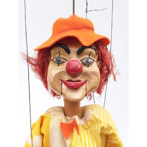 May include: A vintage clown puppet with a painted face, red hair, and an orange felt hat. The puppet wears a yellow and white striped shirt with an orange bow tie. Black strings are attached for manipulation.
