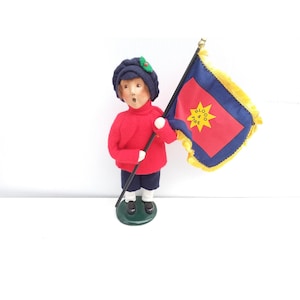 May include: A figurine of a child in a red sweater, navy blue shorts, and a navy blue hat with a green accent. The child holds a flag with a red center, blue border, and gold fringe. The flag has a yellow star and the words "BLOOD & FIRE".