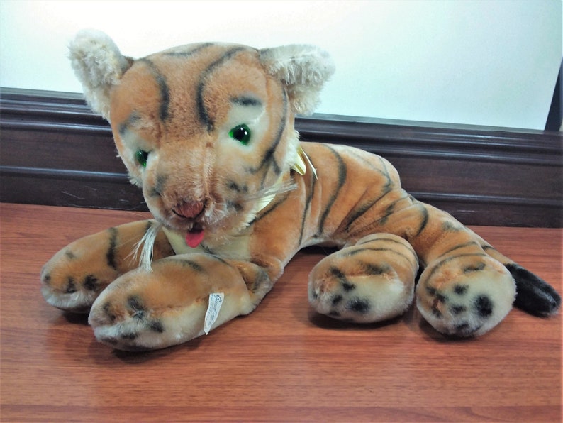 May include: A plush tiger toy with brown and black stripes, green eyes, and a pink tongue sticking out. The toy is lying on a wooden surface.