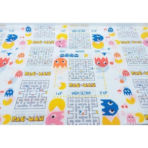 May include: White fabric with a colorful Pac-Man pattern. The design includes mazes, Pac-Man characters, ghosts, and text elements like "PAC-MAN", "HIGH SCORE", and "2 UP". The fabric is suitable for various crafting projects.