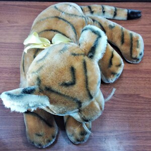 May include: A plush tiger toy with brown and black stripes. The toy has a yellow bow tied around its neck and is lying on a wooden surface.