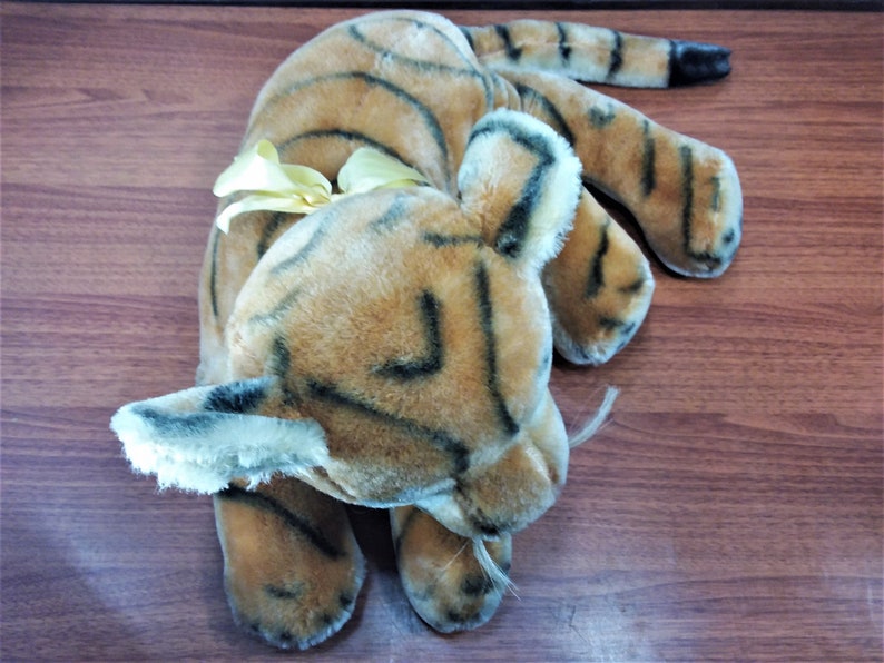 May include: A plush tiger toy with brown and black stripes. The toy has a yellow bow tied around its neck and is lying on a wooden surface.
