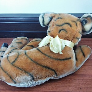 May include: A plush tiger toy with brown and black stripes, lying on its side with its head turned to the right. The tiger has a yellow satin bow tied around its neck.