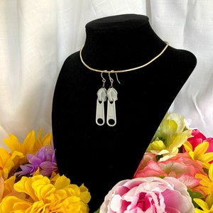 Op de afbeelding: Een paar zilveren rits-oorbellen. De oorbellen hangen aan de nek van een zwart mannequin. Het mannequin is omringd door kleurrijke bloemen.