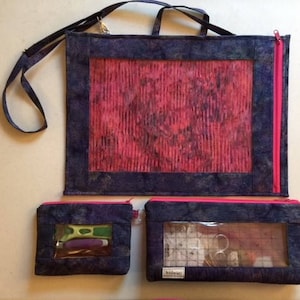 May include: Three zippered bags with clear vinyl windows. The largest bag has a pink and black patterned fabric and a black strap. The two smaller bags have a blue and black patterned fabric. One of the smaller bags has a pink zipper and the other has a black zipper.