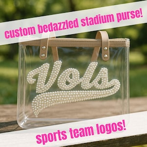 Op de afbeelding: Transparante stadiontas met bruine rand en handvatten. Het woord "Vols" is met witte parels op de voorkant gespeld. De tekst "custom bedazzled stadium purse!" en "sports team logos!" worden in roze weergegeven.