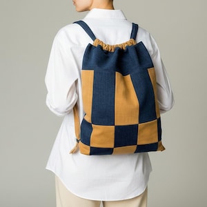 Könnte beinhalten: Ein Patchwork-Rucksack in einem Schachbrettmuster aus Marineblau und Beige. Die Kordelzugtasche hat verstellbare Träger und einen beigefarbenen Kordelzugverschluss. Die Tasche wird über einem weißen Hemd getragen.