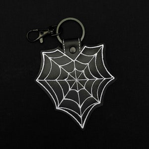 May include: Black leather keychain with a white embroidered spiderweb design. The keychain has a black metal clasp and a key ring. The spiderweb is a classic Halloween or gothic motif.