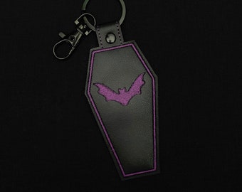 May include: Black coffin-shaped keychain with a purple bat design. The keychain has a black metal clasp and key ring. The bat and coffin are outlined in purple stitching. The keychain is made of a leather-like material.