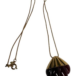 Puede incluir: Un collar de cadena en tono dorado con un colgante en forma de concha estilizada. La parte superior del colgante es de metal dorado liso, mientras que la sección inferior es de un material translúcido de color rojo oscuro con estrías.