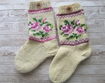 May include: A pair of cream-colored knitted socks with a floral design. The socks feature pink and green roses, with a pink and purple border at the top. Each sock has a wooden tag with a number on it.