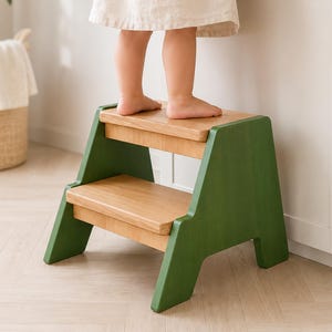 May include: A two-step stool featuring natural light wood treads and solid green side panels. The structure has a wide base for stability with two horizontal steps, providing a functional elevation tool constructed from wood with a painted finish.