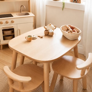 May include: A light-coloured wooden play table with two matching chairs, featuring a toy tea set with a teapot, cups, and a woven basket filled with wooden play food items, positioned near a wooden toy kitchen unit with a sink and stove.