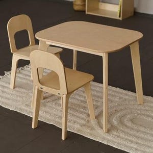 Pode incluir: Uma mesa e duas cadeiras infantis de madeira clara estão sobre um tapete creme. A mesa tem um tampo retangular com cantos arredondados. As cadeiras têm um encosto curvo com um recorte. Uma estante e um berço estão ao fundo.