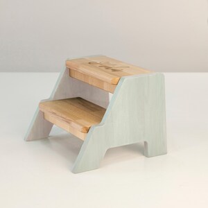 A small, two-tiered wooden step stool with a light-colored wooden top and a darker wooden bottom.