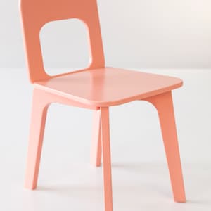 May include: A small, peach-colored wooden chair with a square seat and a backrest featuring a rectangular cutout. The chair has four straight legs and a simple, modern design. The chair is suitable for children.