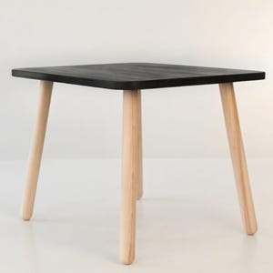 Peut inclure: Petite table carrée avec un plateau noir et quatre pieds en bois clair. La table mesure environ 50 cm de large et 46 cm de haut, idéale pour une chambre d'enfant ou comme table d'appoint.