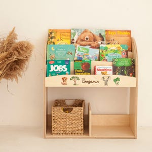 May include: A light-colored wooden bookshelf with multiple children's books. The bookshelf has two shelves, with a woven basket on the bottom shelf. The top shelf displays books with titles like "Jobs" and "Town". The name "Benjamin" is written on the front.