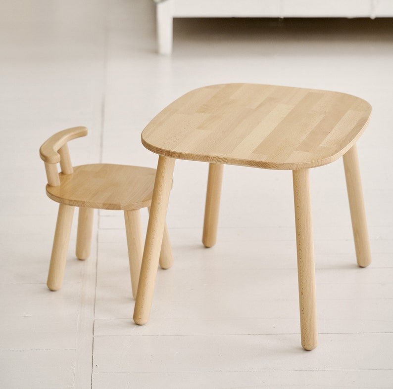 K&ouml;nnte beinhalten: Ein kleines, helles Tisch- und Stuhlset aus Holz. Der Tisch hat eine abgerundete quadratische Oberseite und vier Beine. Der Stuhl hat eine gebogene R&uuml;ckenlehne und vier Beine. Das Set ist aus hellem Holz gefertigt.