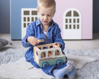 May include: A child playing with a wooden toy bus. The bus is light blue with red and green accents and blue wheels. The child is wearing a denim shirt and blue jeans. Two large, colorful house-shaped structures are in the background.