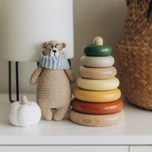 Könnte beinhalten: Ein Holz-Stapelspielzeug mit bunten Ringen in den Farben Grün, Beige, Gelb, Orange und Braun. Das Spielzeug befindet sich auf einer weißen Oberfläche neben einem gehäkelten Bären mit Brille und einem blauen Schal.