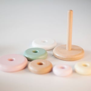 May include: A wooden stacking toy with a natural wood base and post, surrounded by six colorful, round rings in pastel shades of pink, green, beige, and yellow. The toy is designed for children and promotes hand-eye coordination.