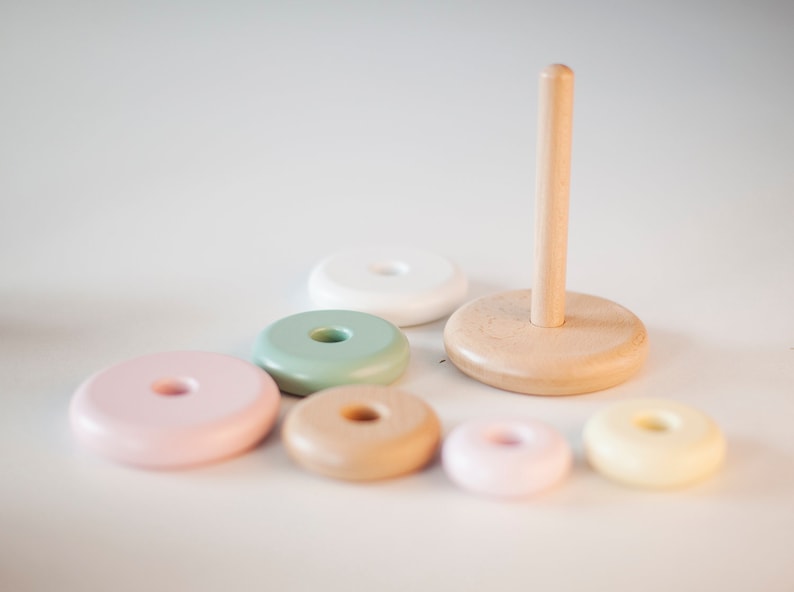 May include: A wooden stacking toy with a natural wood base and post, surrounded by six colorful, round rings in pastel shades of pink, green, beige, and yellow. The toy is designed for children and promotes hand-eye coordination.