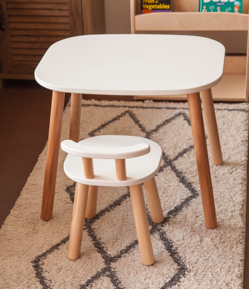 K&ouml;nnte beinhalten: Ein wei&szlig;er Kindertisch mit abgerundeten Ecken und vier Holzbeinen. Ein passender wei&szlig;er Stuhl mit geschwungener R&uuml;ckenlehne und Holzbeinen steht vor dem Tisch. Tisch und Stuhl stehen auf einem gemusterten Teppich.