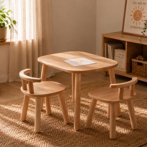 May include: A light-colored wooden children's table and two chairs set. The table has a square top with rounded corners. The chairs have curved backs. The set is placed on a woven rug. A framed print with a sun illustration is visible.