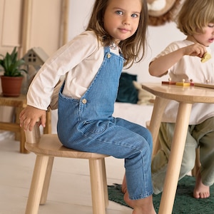 K&ouml;nnte beinhalten: Ein junges Kind in einer blauen Cord-Latzhose und einem wei&szlig;en Langarmshirt sitzt auf einem kleinen Holzstuhl. Das Kind befindet sich in einem Raum mit einem Holztisch und einem anderen Kind. Eine L&ouml;wenkopfdekoration befindet sich an der Wand.