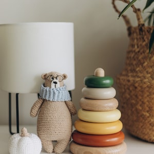 May include: A crocheted bear wearing glasses and a blue scarf sits next to a wooden stacking ring toy with colorful rings. A small white pumpkin and a lamp are also in the image. A woven basket is in the background.