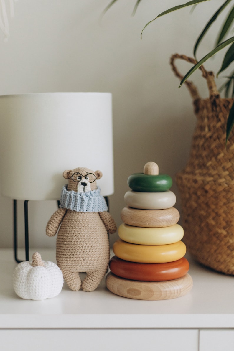 May include: A crocheted bear wearing glasses and a blue scarf sits next to a wooden stacking ring toy with colorful rings. A small white pumpkin and a lamp are also in the image. A woven basket is in the background.