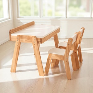 May include: A children's furniture set consisting of a rectangular table with a white top and light wood legs, accompanied by two matching chairs featuring white seats and light wood frames, all constructed with visible metal hardware fasteners.