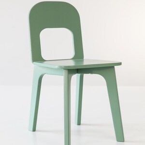 May include: A solid, sage green wooden chair with a square seat and a backrest featuring a rectangular cutout. The chair has four straight legs and a curved back, suitable for a child's room or a minimalist space.