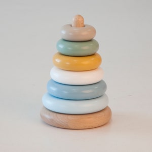 Puede incluir: Un juguete de apilar de madera con seis anillos coloridos. Los anillos son en tonos de azul, verde, amarillo y beige. El juguete está diseñado para bebés y niños pequeños.