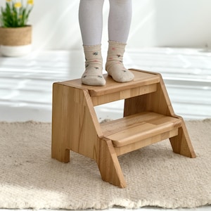 A child's feet standing on a small wooden step stool in a room with a rug and a potted plant.