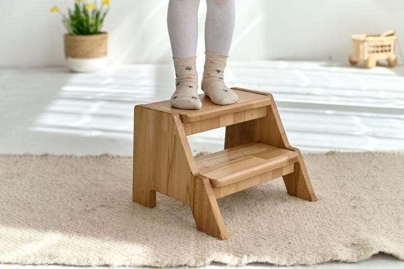 A child's feet standing on a small wooden step stool in a room with a rug and a potted plant.