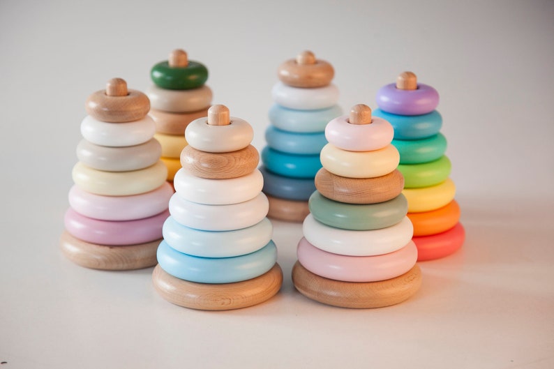 May include: Six wooden stacking ring toys in pastel colors. Each toy has a wooden base, rings in various colors, and a wooden top. The colors include pink, blue, green, yellow, and purple. The toys are arranged on a white surface.