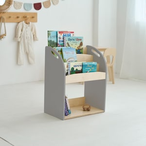 May include: A grey wooden bookcase with three shelves, displaying a selection of colourful children's books. The bookcase has a simple, modern design with curved handles. The background includes a white wall and a wooden coat rack.