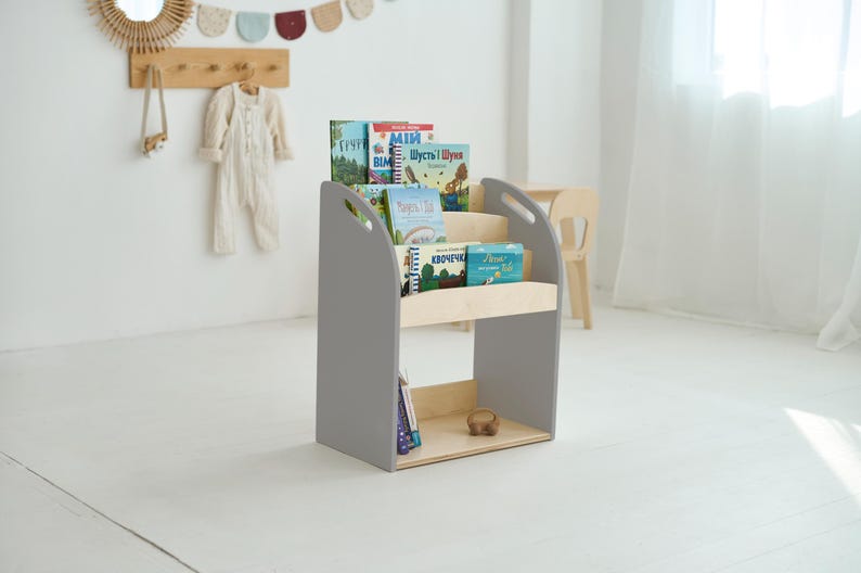 May include: A grey wooden bookcase with three shelves, displaying a selection of colourful children's books. The bookcase has a simple, modern design with curved handles. The background includes a white wall and a wooden coat rack.