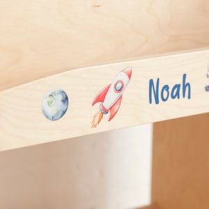 May include: A light-coloured wooden surface featuring a watercolour illustration of a rocket ship and planet Earth. The name "Noah" is written in blue script. The rocket is red and white with orange flames.