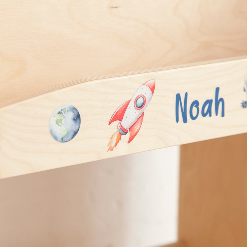 May include: A light-coloured wooden surface featuring a watercolour illustration of a rocket ship and planet Earth. The name "Noah" is written in blue script. The rocket is red and white with orange flames.