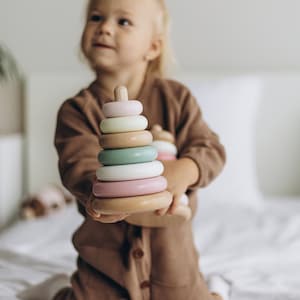 May include: A wooden stacking toy with pastel-colored rings. The toy has a light pink, white, green, and light pink ring. The base and top are made of natural wood. The toy is held by a child wearing a brown jumpsuit.