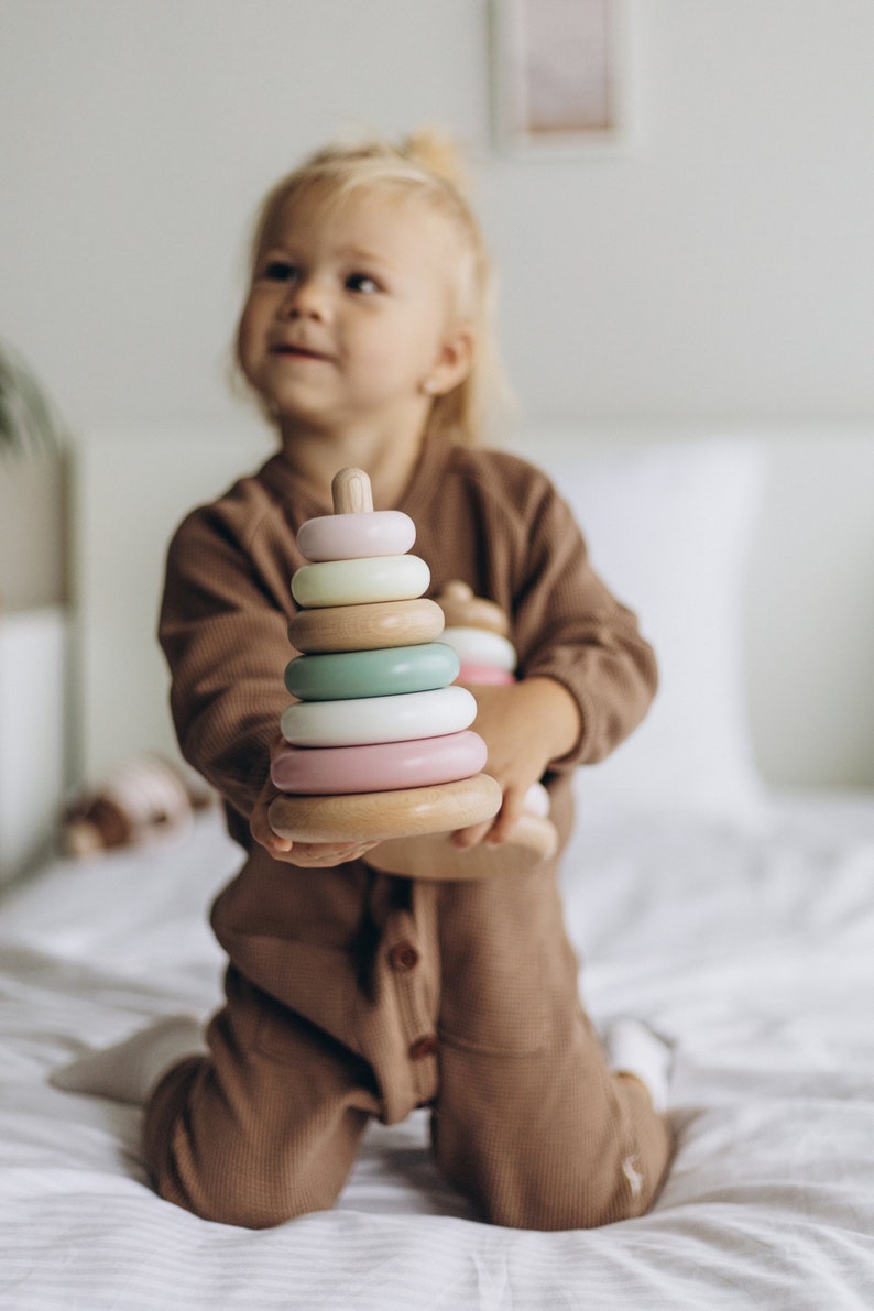 May include: A wooden stacking toy with pastel-colored rings. The toy has a light pink, white, green, and light pink ring. The base and top are made of natural wood. The toy is held by a child wearing a brown jumpsuit.