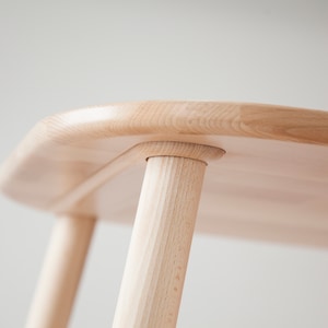 K&ouml;nnte beinhalten: Nahaufnahme eines hellen Holztisches. Der Tisch hat eine abgerundete Oberseite und vier zylindrische Beine. Die Beine sind mit einer Zapfen- und Schlitzverbindung an der Tischplatte befestigt. Die Holzmaserung ist sichtbar.