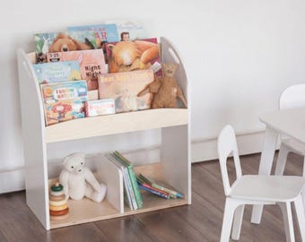 May include: A white wooden bookshelf with a natural wood shelf and base, filled with children's books. A teddy bear and a wooden stacking toy sit on the bottom shelf. A white chair and table are in the background.
