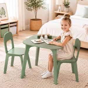 May include: A sage green children's table and two matching chairs set. The table has a square top with rounded corners and four angled legs. Each chair features a curved backrest with a central cutout. The furniture is made of smooth wood.