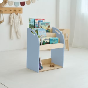 May include: A pale blue wooden bookcase with three shelves, displaying various colourful children's books. The bookcase has a light wood shelf and base. The design includes handles on the sides. The room has a white wall and floor.